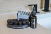 Beard grooming set with brush, comb, and bottle on a bathroom counter.