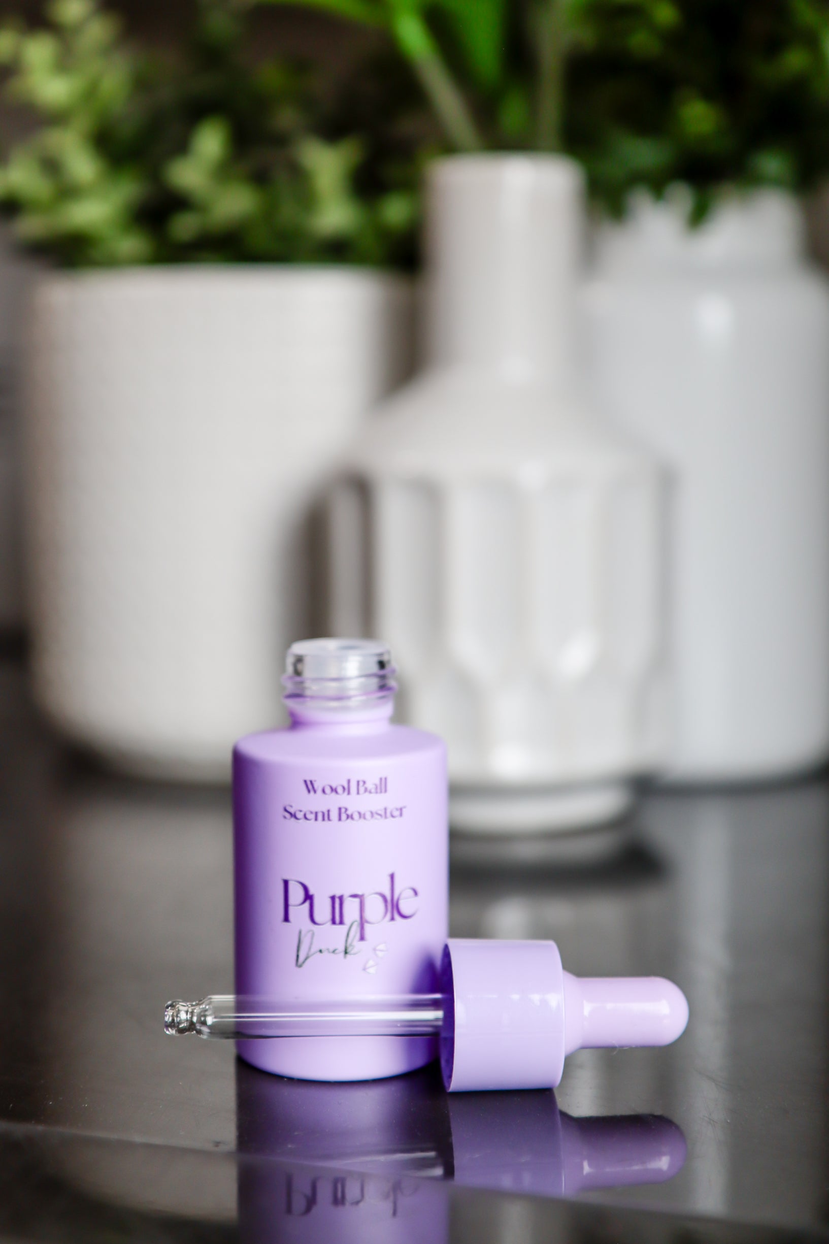 Purple bottle of Scent Boosters showing dropper applicator on a reflective surface with white vases and greenery in the background.