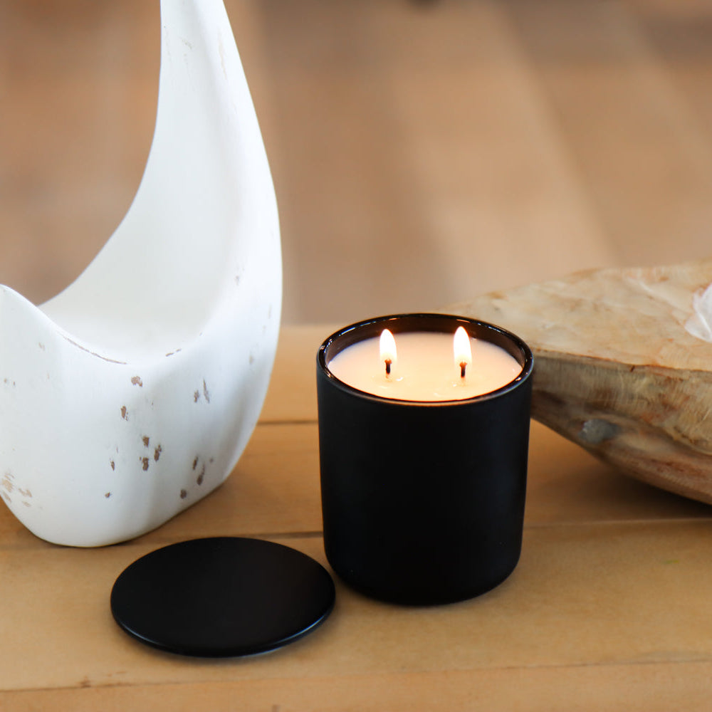 A Purple Duck Men's black candle with a lit flame, placed on a wooden surface, with decorative items in the background.