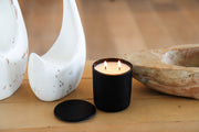 A Purple Duck Men's black candle with a lit flame, placed on a wooden surface, with decorative items in the background.