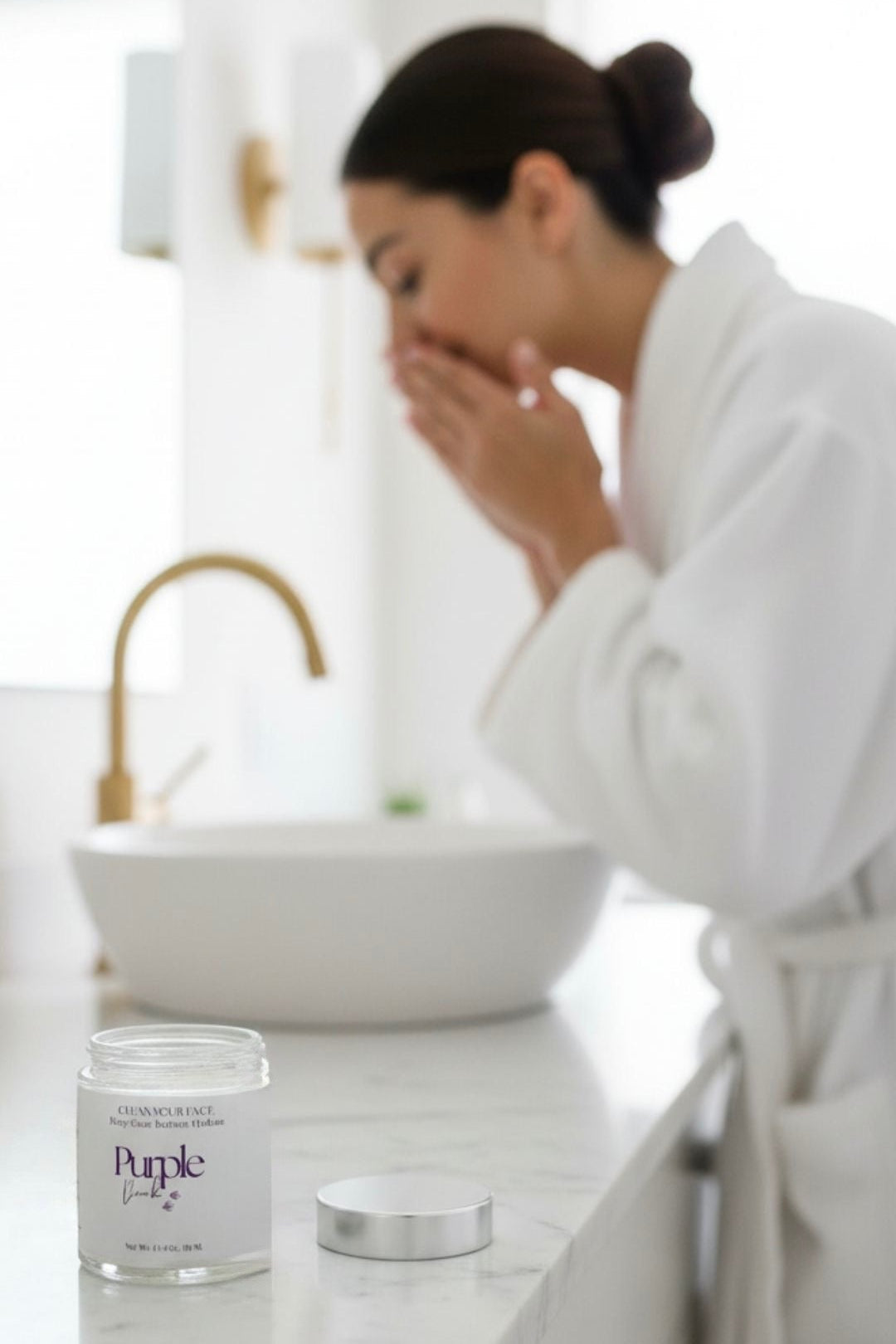 Woman in a bathrobe applying facial cleanser to her face with the jar of facial cleanser on the counter