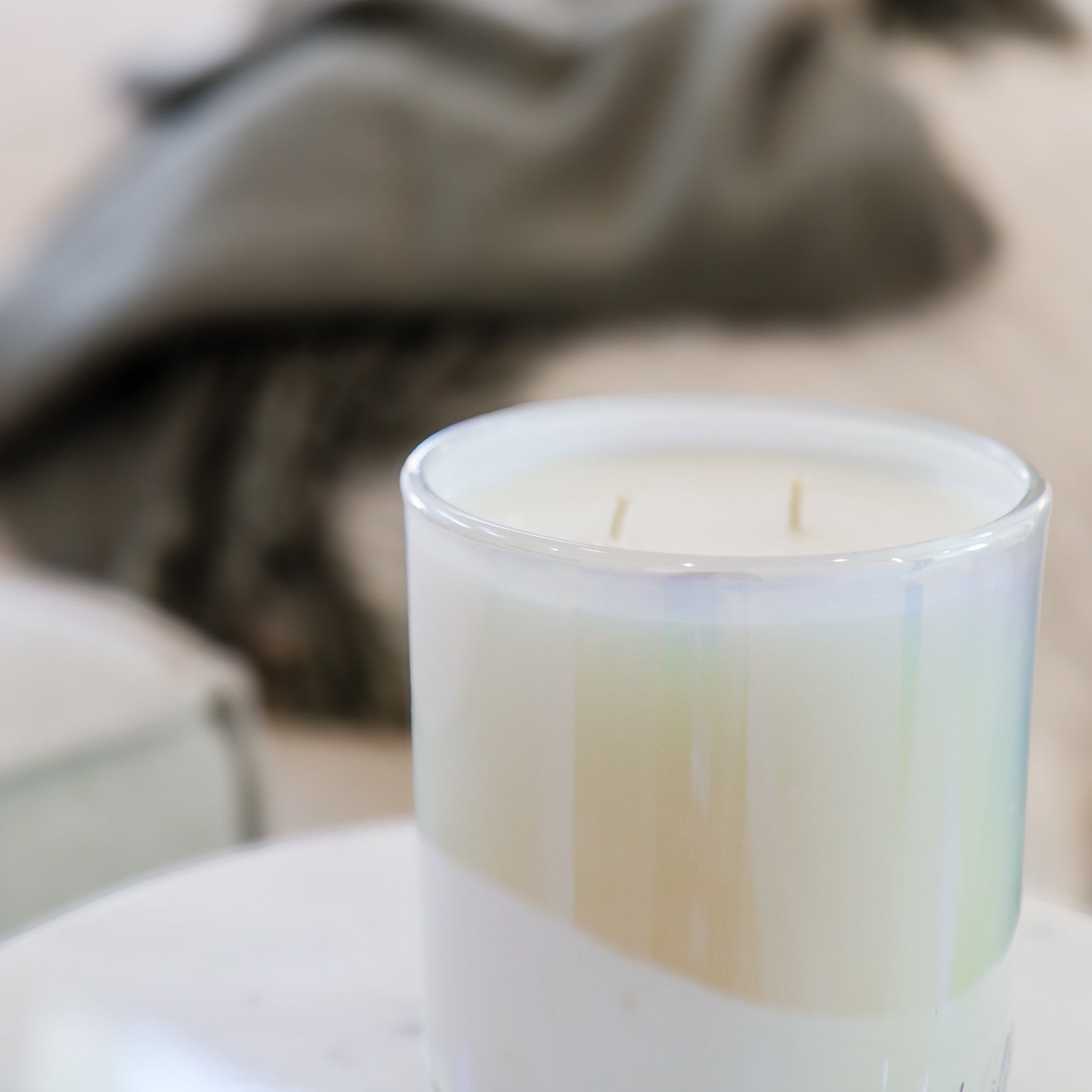 Candle in a glass container on a table with a blurred background showing a couch and plant.