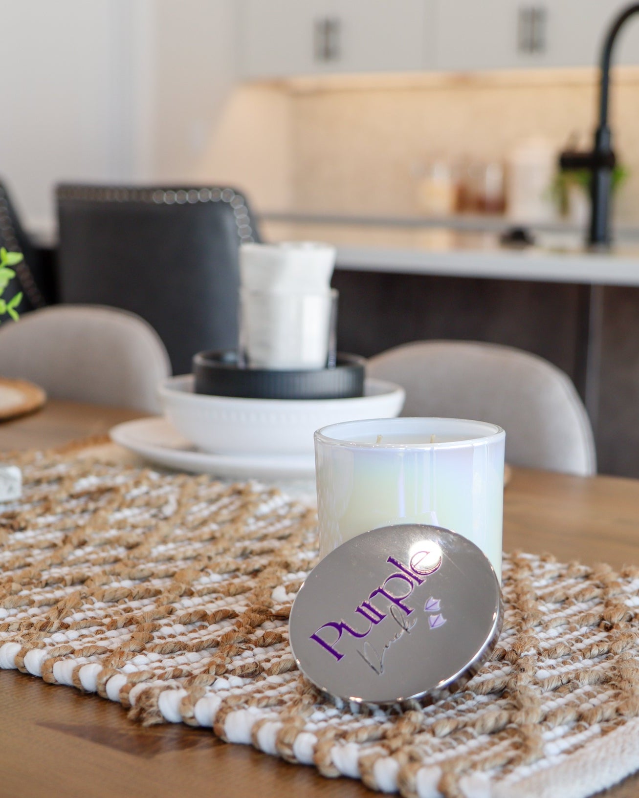 A white, two-wick coconut blend wax candle placed inside a clear, foggy vessel. The candle is set on a wooden surface with a decorative mat and is accompanied by a decorative plant and a white candle holder in the background.