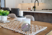A white, two-wick coconut blend wax candle placed inside a clear, foggy vessel. The candle is set on a wooden surface with a decorative mat and is accompanied by a decorative plant and a white candle holder in the background.