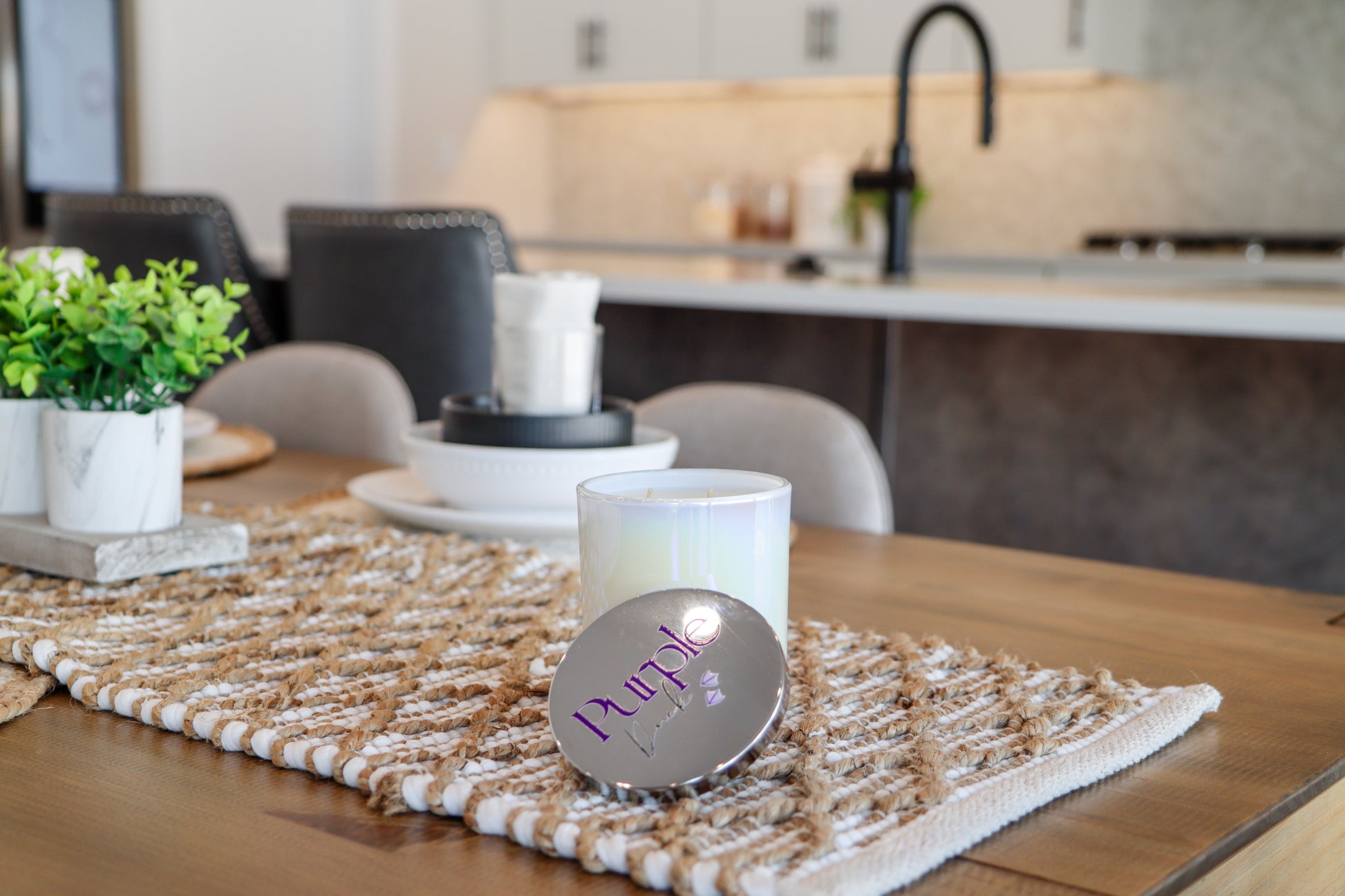 A white, two-wick coconut blend wax candle placed inside a clear, foggy vessel. The candle is set on a wooden surface with a decorative mat and is accompanied by a decorative plant and a white candle holder in the background.