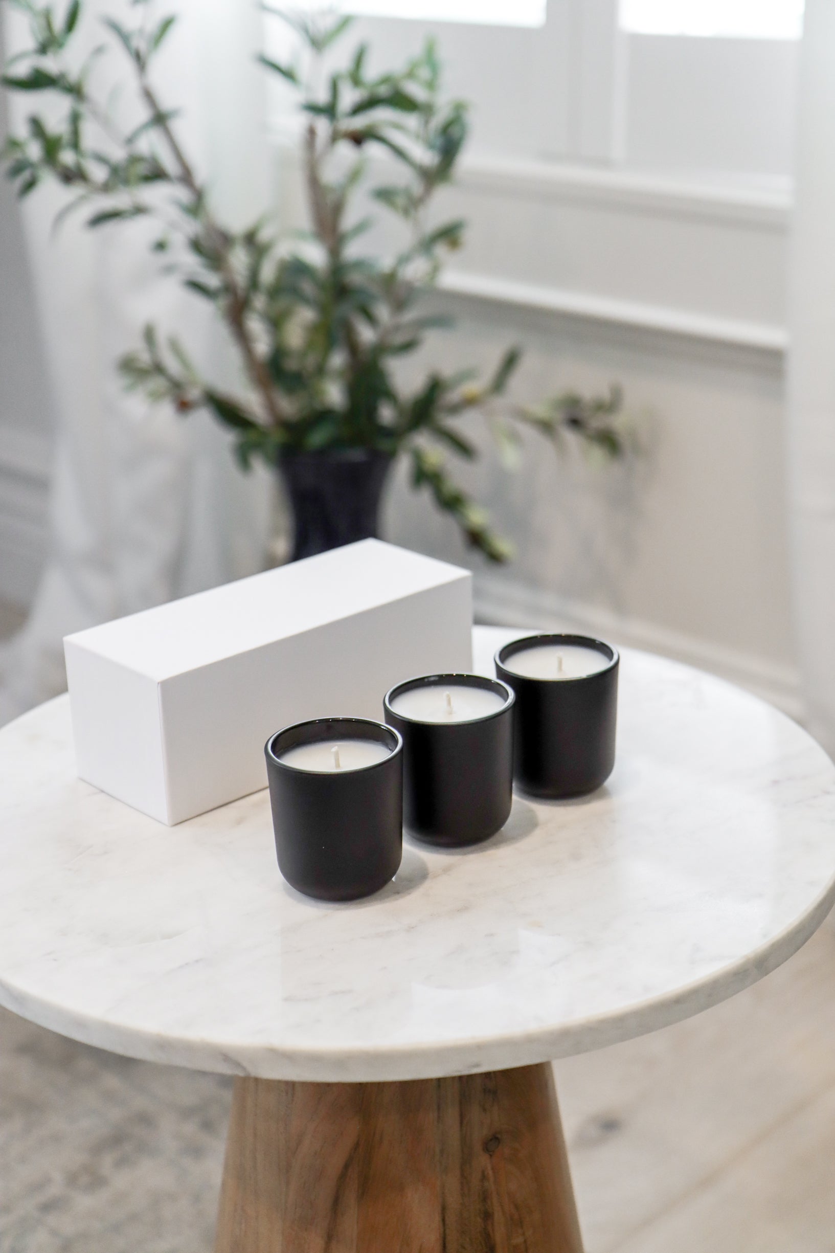 Three black candles on a marble table with a white box and a plant in the background.