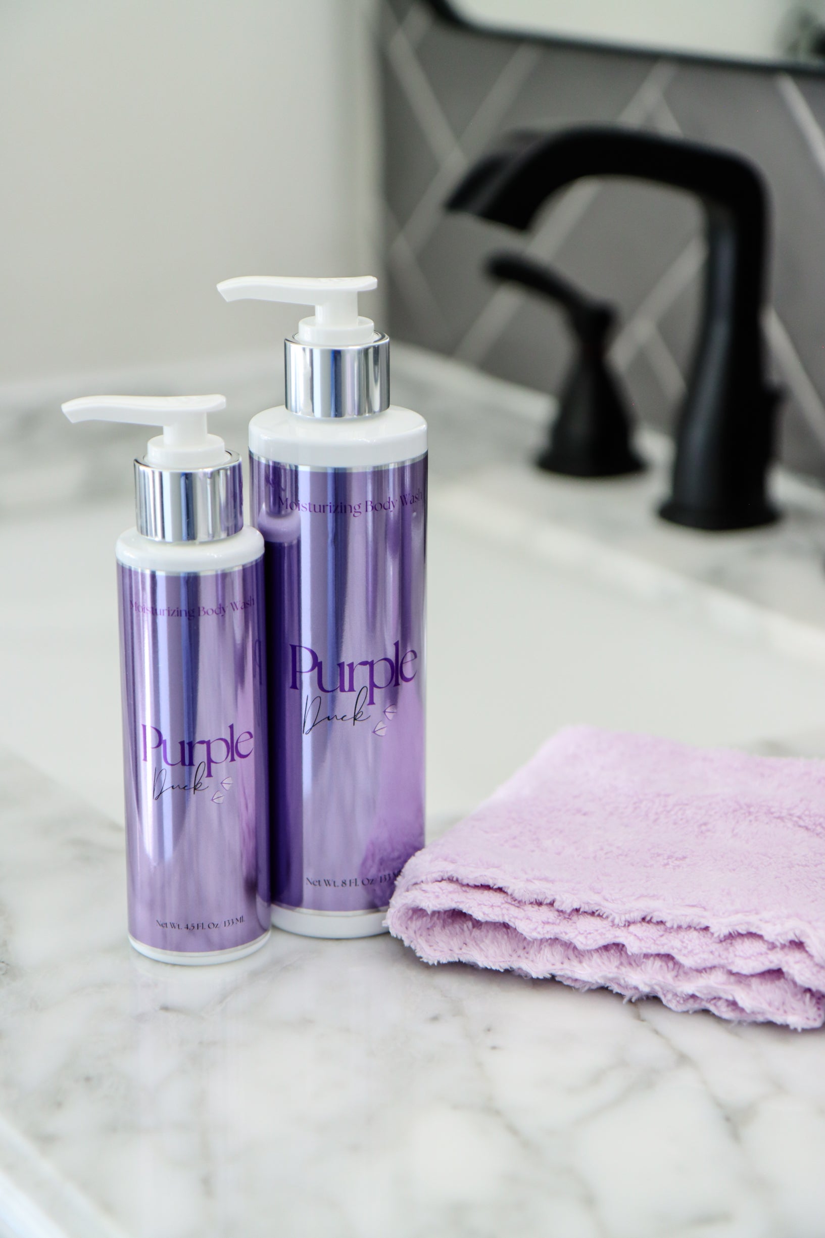 Both sizes of body wash, 4.5 oz and 8oz on a bathroom counter with a towel and faucet in the background.