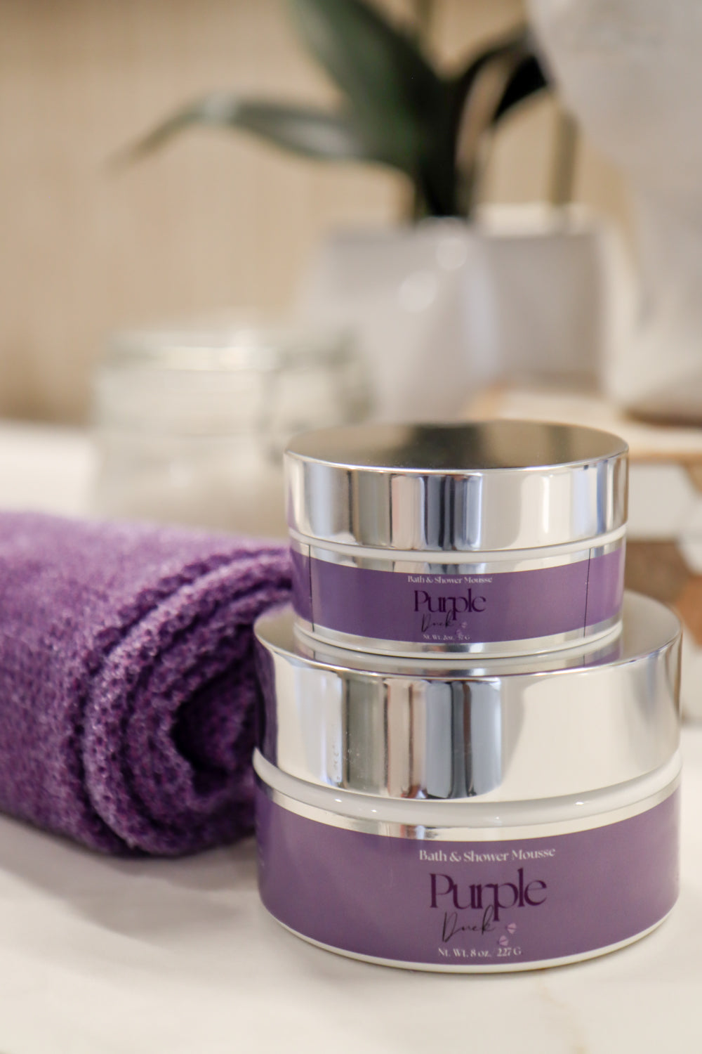 A stack of purple and silver colored bath and shower mousse containers with a purple towel and plant in the background.