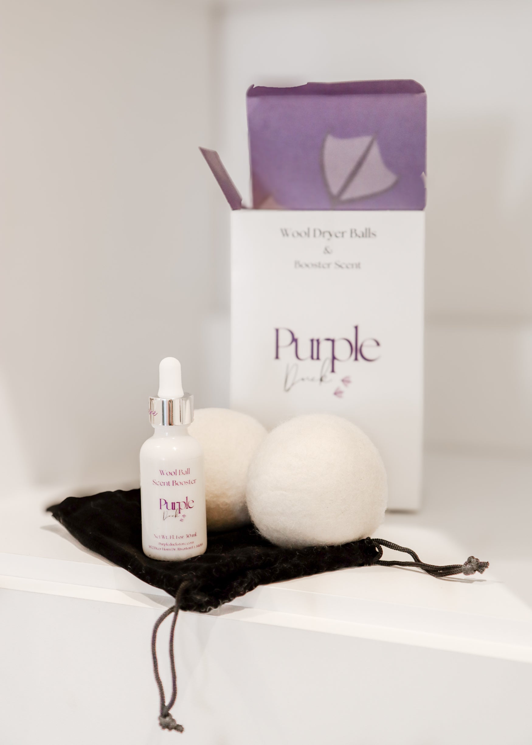A set of white wool dryer balls next to a box of Purple Duck brand dryer balls with a small bottle of fragrance oil, all displayed on a dark cloth surface.